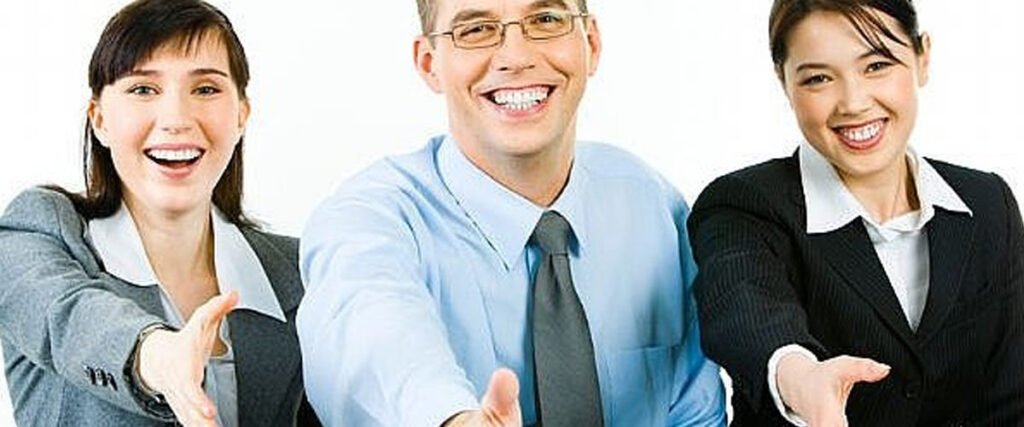 Three smiling professionals, two women and one man, all dressed in business attire, extend their right hands forward as if to greet or shake hands. The plain white background gives a clean and focused look on the individuals, resembling a confident team ready for a PowerPoint presentation.