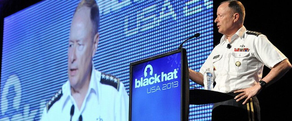 A man in a military uniform speaks at a podium with a "Black Hat USA 2013" sign while his large projected slide template appears on a screen behind him. He holds a water bottle and stands in front of a blue dotted background.