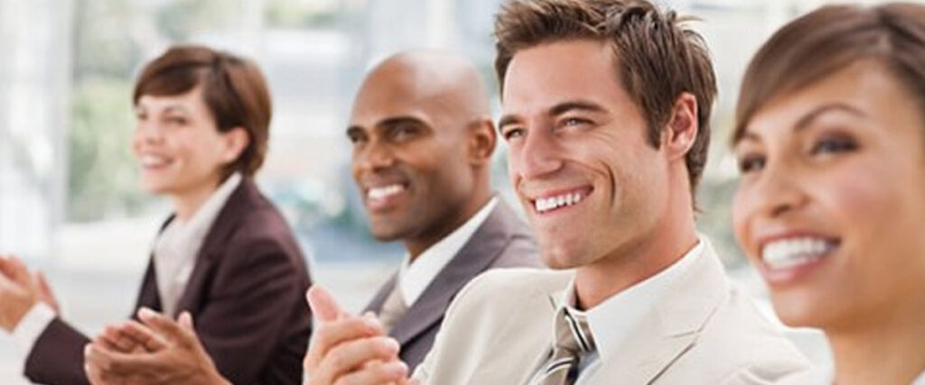 A diverse group of four people in business attire sits in a row, smiling and clapping. They appear to be attending a professional event or meeting, with a PowerPoint slide projected in the background, suggesting an indoor setting with bright, natural light.