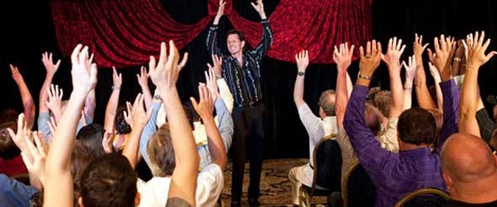 A man stands on stage with hands raised in the air, facing an audience who have also raised their hands. The backdrop is adorned with red curtains. The audience is seated in rows of chairs, and the atmosphere appears energetic and lively, as if they are engaged in an interactive presentation.