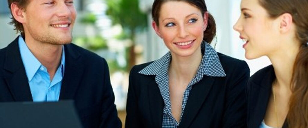 Three people in business attire are seated and engaged in a lively conversation. One man and two women are smiling, with the woman on the right speaking. There's a laptop in front of the man displaying a pitch deck. The background is an office-like setting with plants and bright lighting.