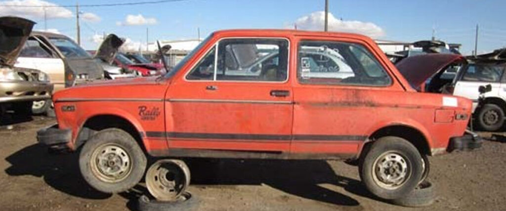 An old, red, two-door car missing its front left wheel is parked in a scrapyard, with other vehicles and car parts visible in the background under a partially cloudy sky. The car is supported by two tire rims on that side. This scene could serve as an interesting element for a slide template or presentation backdrop.