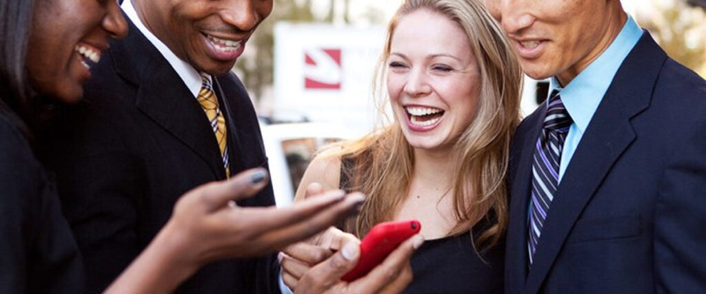 A group of four people, dressed in business attire, are standing close together and smiling while looking at something on a red smartphone. They appear to be outdoors on a sunny day, possibly reviewing a pitch deck or PowerPoint slide.