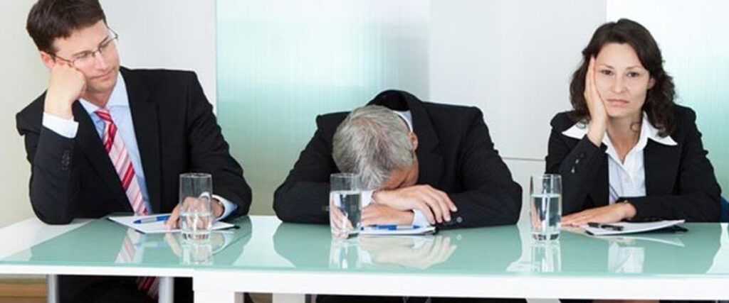 Three professionals in business attire are seated at a glass-top table with glasses of water and notepads. They look bored; the man in the middle has his head on the table, while the man and woman on either side rest their heads on their hands. Clearly, this presentation pitch deck isn't engaging them.