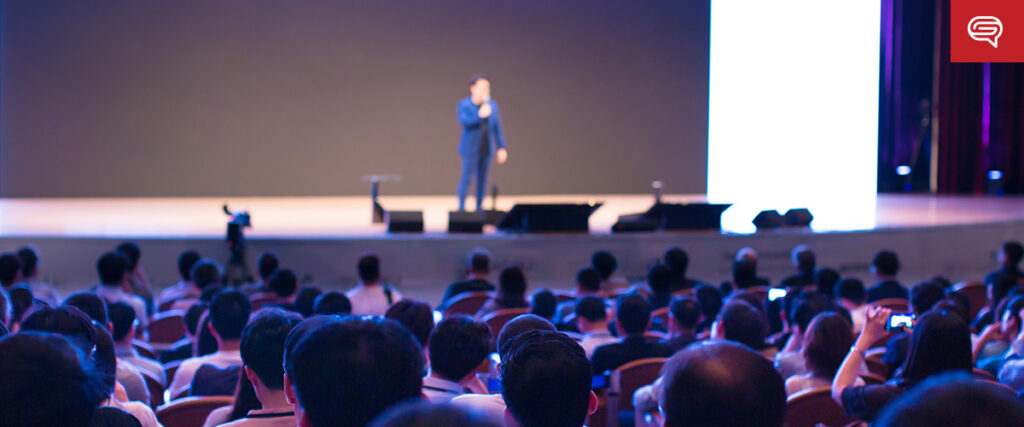 A speaker dressed in a blue suit stands on stage addressing an audience seated in an auditorium. The stage is lit with bright lights, and there’s a large, illuminated slide behind the speaker. The audience members are focused, some holding up phones to record.