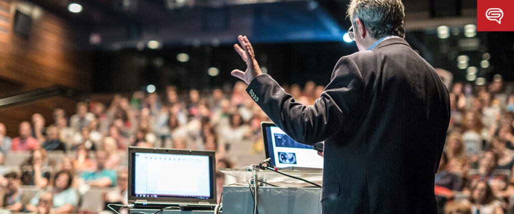 A person in a suit speaks at a podium, gesturing with one hand, while addressing a large audience in a lecture hall. A laptop and projector are set up on the podium, displaying a PowerPoint slide template. The audience is seated and focused on the speaker.