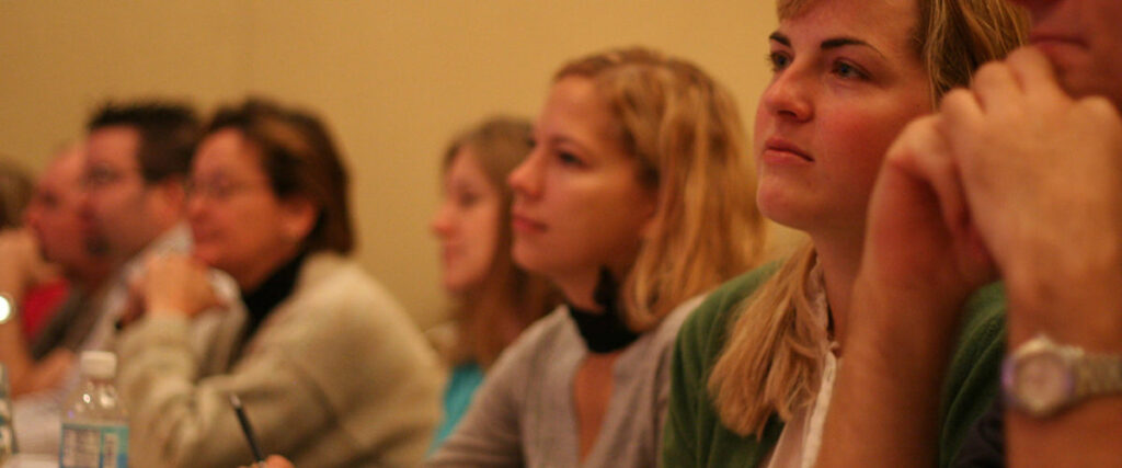 A group of people sit at a table, attentively listening and taking notes during a presentation. Some individuals hold pens and notebooks, while others rest their hands on their faces. The background is softly blurred, focusing on the participants' engaged expressions as they follow along with the pitch deck.