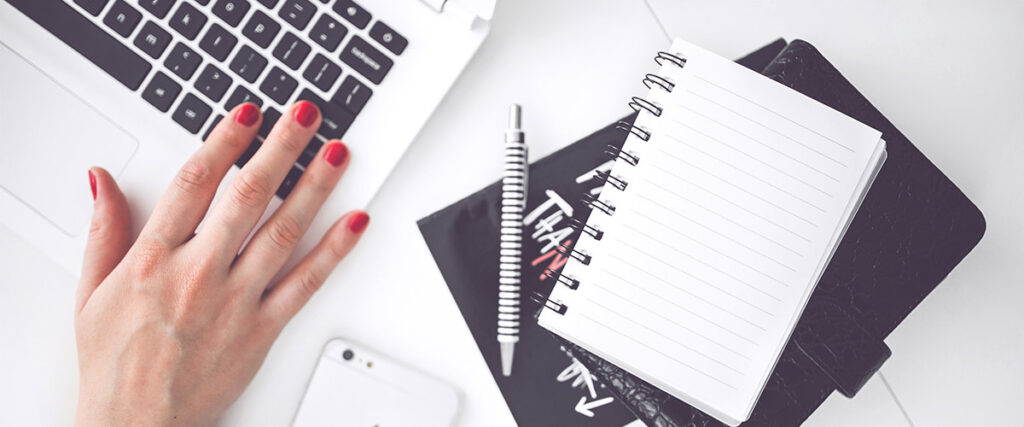 A person's hand with red nail polish rests on a laptop keyboard. Nearby are a blank spiral notebook, a pen, and a closed planner. An iPhone is partially visible on the left side of the workspace, ready for notes or updates during a pitch deck creation.