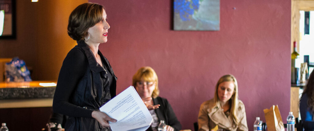 A person with short hair is standing indoors, holding papers and presenting a pitch deck. Three people are seated in the background, listening attentively. The room has maroon walls with a painting and modern decor. Water bottles and papers are on the tables.