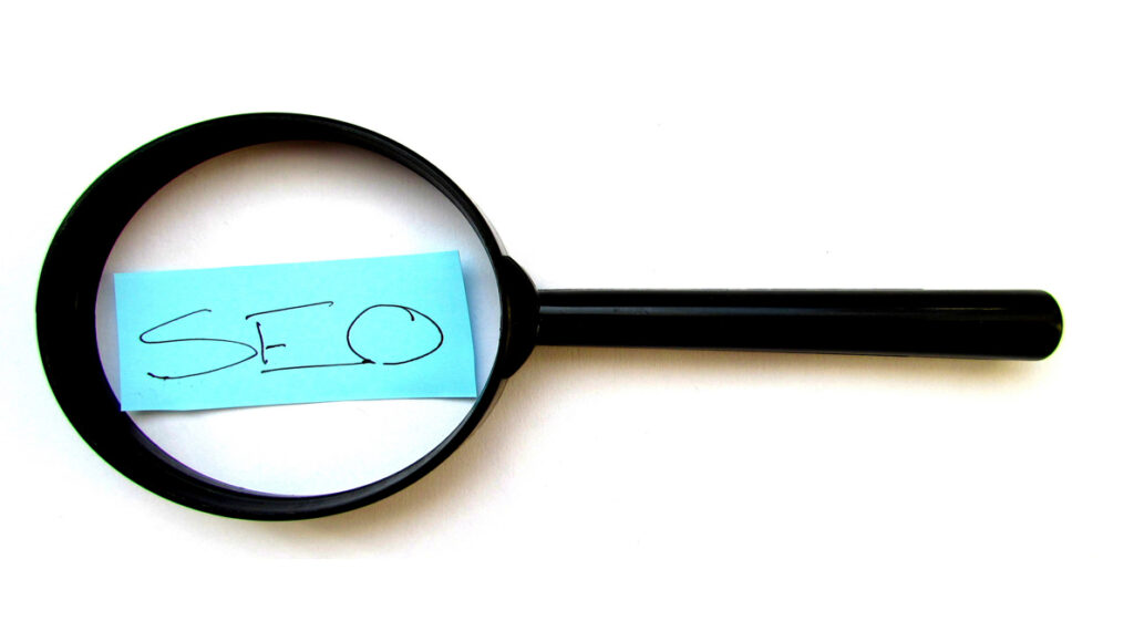 A black magnifying glass with a light blue sticky note on its lens displaying the handwritten letters "SEO" in black ink. The magnifying glass is positioned on a white background, making it perfect for using in your next slide template or pitch deck.