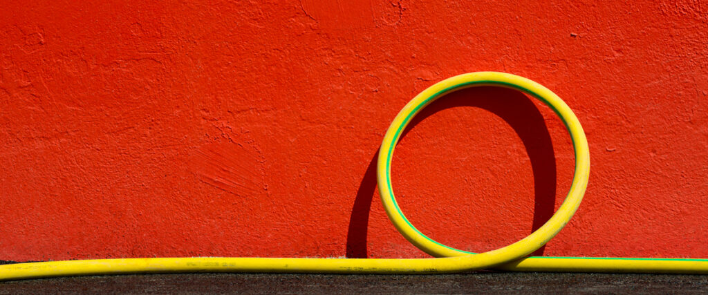 A yellow garden hose is coiled into a circle against a vibrant red stucco wall, creating a contrasting and visually appealing composition. The hose lies partially on a dark surface beneath the wall, making for an eye-catching background in any presentation.