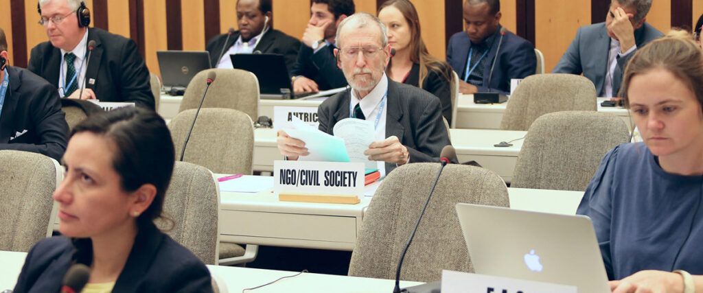 A group of people are sitting in a conference room, each at their own desk with laptops and documents. In the center, an older man with glasses reads a paper labeled "NGO Civil Society." Other attendees are focused on their pitch decks or listening attentively.