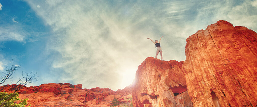 A person stands triumphantly with arms raised on top of a tall, rocky cliff under a bright sky with wispy clouds. The scene is bathed in warm sunlight, highlighting the vibrant red and orange hues of the rock formation; a perfect visual for an inspiring slide in your next pitch deck.