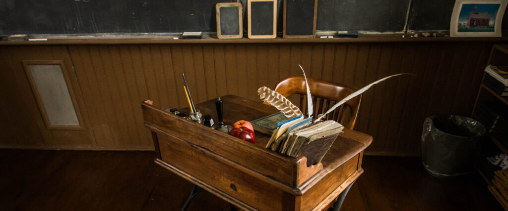 A wooden school desk with an ink bottle, quills, old books, and an apple on top. Behind the desk is a wooden chair and a chalkboard with framed paper remnants and a small picture framed. The setting appears to be an old-fashioned classroom, reminiscent of classic presentation materials.