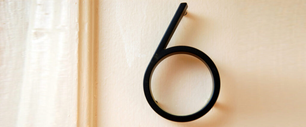 A black number six is mounted on a light-colored wall, resembling an element you'd find in a modern and sleek PowerPoint slide with its hollow circular bottom.