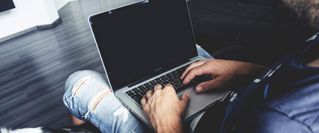 A person wearing ripped jeans and a dark shirt types on a laptop while sitting. The background features a minimalist room with wooden flooring and a dark sofa, possibly working on a PowerPoint pitch deck or slide template.