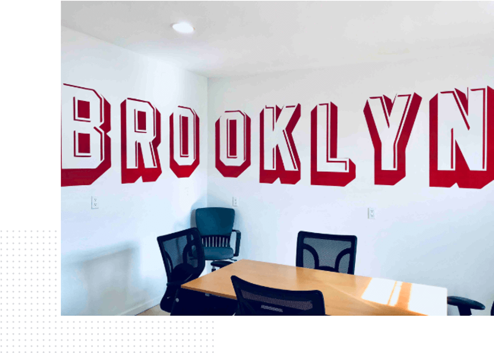 A small conference room with a wooden table and three black mesh chairs is shown. The white wall behind them has the word "BROOKLYN" painted in large, bold red letters with white outlines. An additional teal chair is seen in the corner of the room, perfect for quick presentations or PowerPoint slide reviews.