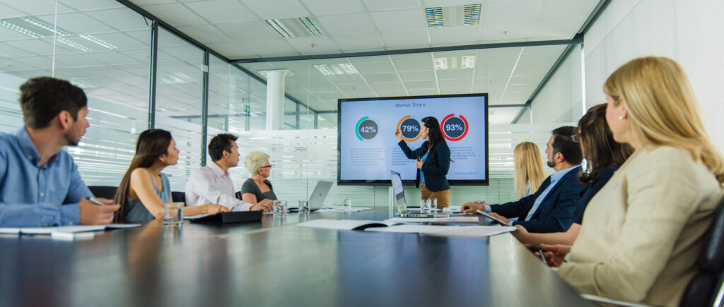 A group of people are seated around a conference table in a modern office, attentively watching a presenter in front of them. The presenter points to a screen displaying colorful pie charts and statistics from their pitch deck, indicating a business presentation or meeting.