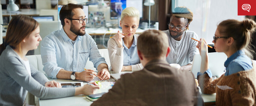 A diverse group of six people is gathered around a table in an office setting having a discussion. They appear engaged, each holding pens and papers. The bright room features large windows in the background. One person is looking thoughtfully at the other participants as they discuss their pitch deck.