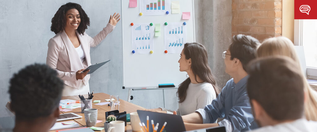 A businesswoman stands smiling and presenting a pitch deck on a whiteboard to a group of five colleagues seated around a table. The charts display various graphs. The setting appears to be a modern office with exposed brick walls.