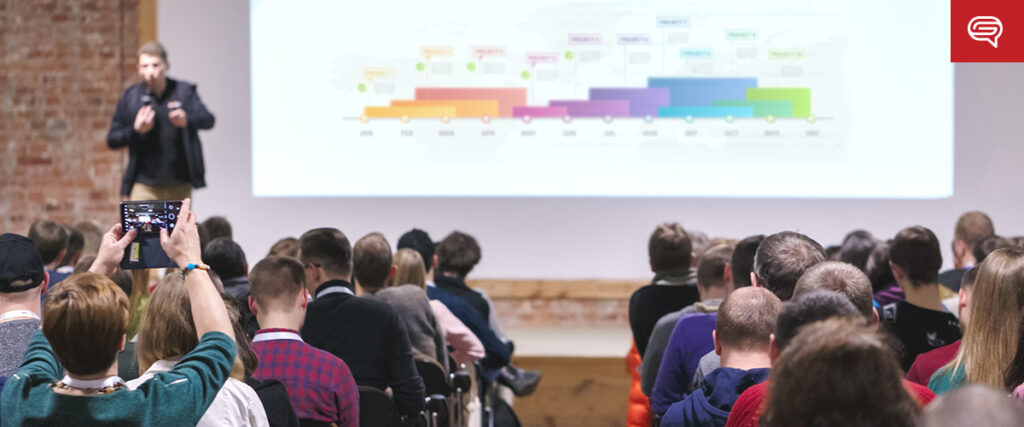 A speaker presents a colorful bar graph slide to an engaged audience in a packed lecture hall. One audience member takes a photo with a smartphone. A red logo is visible in the top right corner of the presentation.