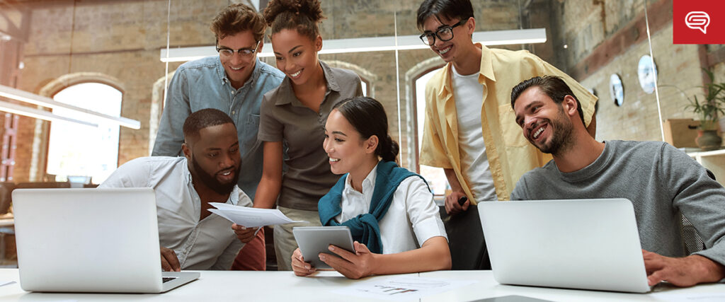 A group of five diverse people is gathered around a table in a modern office setting. They are engaged in a discussion, looking at documents and digital devices, including laptops and a tablet with a PowerPoint slide. All appear focused and collaborative, with a bright, spacious background.