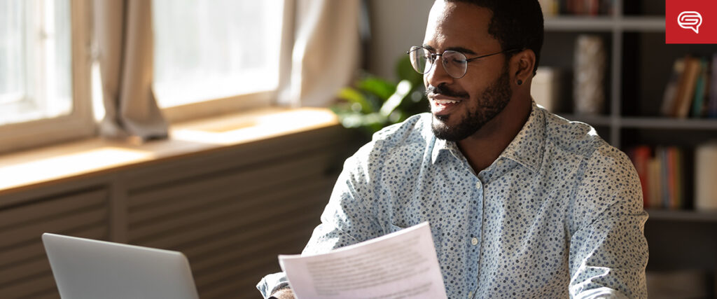 A man wearing glasses and a patterned shirt sits at a desk, holding a document and looking at his laptop with a PowerPoint slide open. Sunlight streams through a nearby window, and a bookshelf is visible in the background.