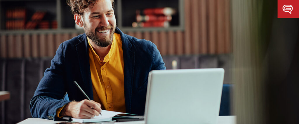 A man with a beard and curly hair, wearing a yellow shirt and dark blazer, sits at a table with an open laptop. He is holding a pen and writing in a notebook, smiling as he looks at the screen. Books are visible on a shelf in the background, possibly preparing for his next PowerPoint presentation.