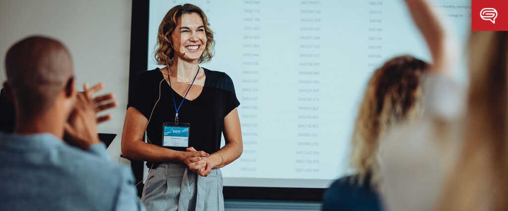 A woman with a microphone headset and name badge smiles while standing in front of a presentation screen displaying a pitch deck. She is engaging with an audience, some of whom are clapping. The background shows a blurred list of dates and text on the slide.