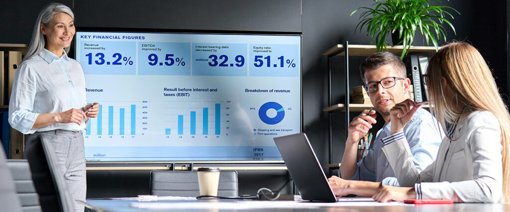 Three business professionals are in a meeting room. A woman stands near a screen displaying graphs and financial figures from her slide presentation. Two people are seated at a table, looking at documents, with one person holding a pen. A plant and shelves are in the background.