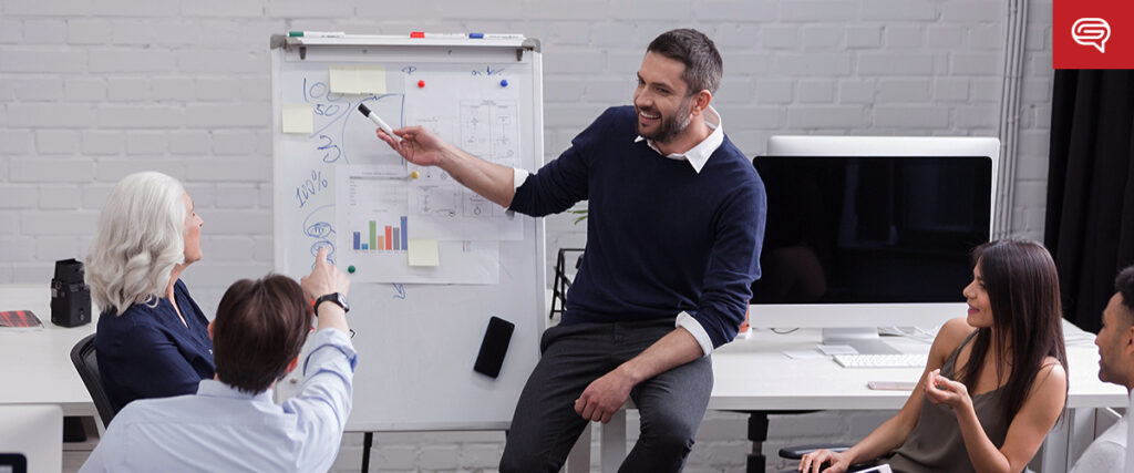 A man with short hair and a beard is smiling and pointing at a slide on a flipchart while sitting on a table. Three people, two women and one man, are seated around him, engaged in the presentation. The background includes a white brick wall and office equipment.