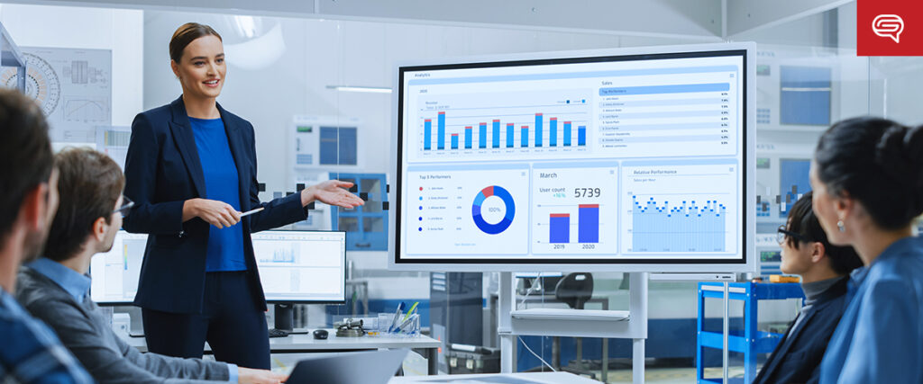 A woman in business attire stands by a large digital screen, holding a presentation pointer. The screen displays various PowerPoint slides with charts, graphs, and data analytics. Four colleagues sit around a table, attentively looking at the presentation in a modern office setting.