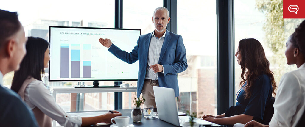 A man in a blue blazer and white shirt is giving a presentation to four people seated around a table. He is pointing at a screen displaying a bar graph and a line chart from his slide template. The room has large windows with natural light coming in.