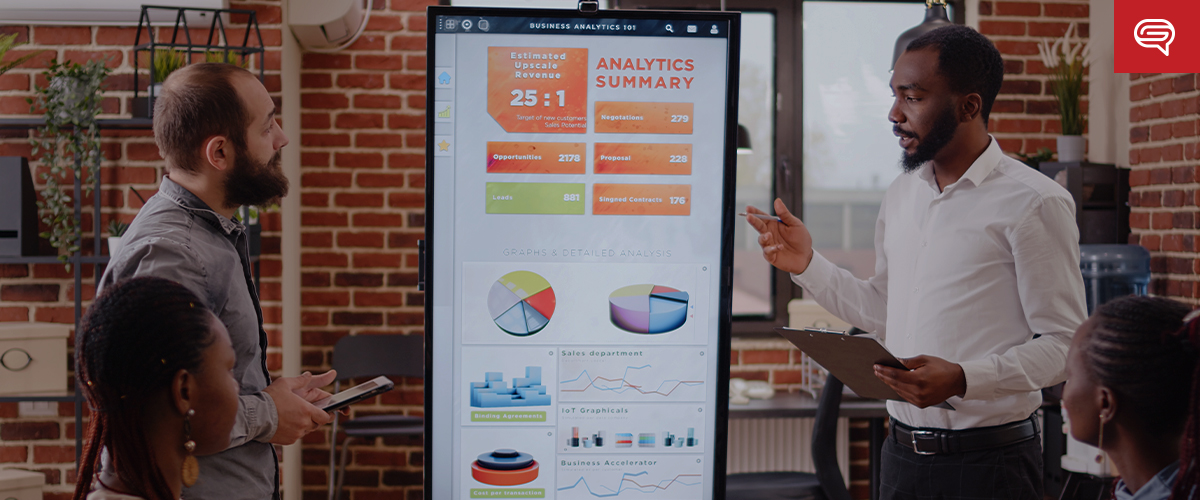 Two people are standing beside a large digital screen displaying an "Analytics Summary." One, holding a tablet, has a beard and is listening. The other, with a clipboard, is gesturing toward the screen. Three people are seated in front, focused intently on the PowerPoint presentation.