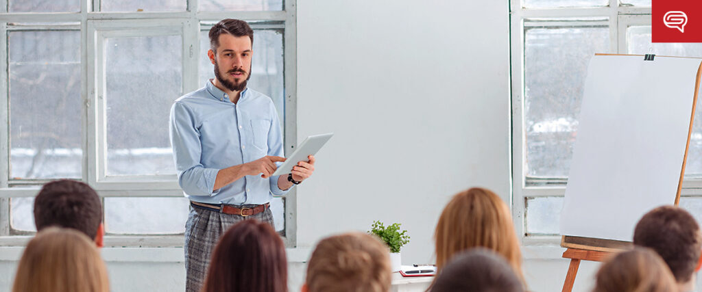 A person with a beard is standing in front of a group, holding a tablet and presenting. They are near a flip chart in a bright room with large windows, using a pitch deck to engage the audience.