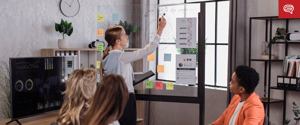 A woman is writing on a clear glass board with colored sticky notes and diagrams, while three colleagues observe attentively. A computer screen displaying charts and a PowerPoint slide is visible to the left. The room has a modern design with plants and shelves in the background.