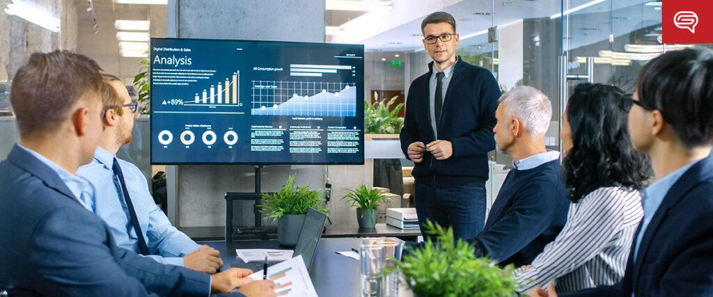 A business meeting in a modern office. Six professionals are seated around a table, listening to a person stand and present data shown on a large screen displaying various PowerPoint slides with charts and graphs. Small plants and books are on the table.