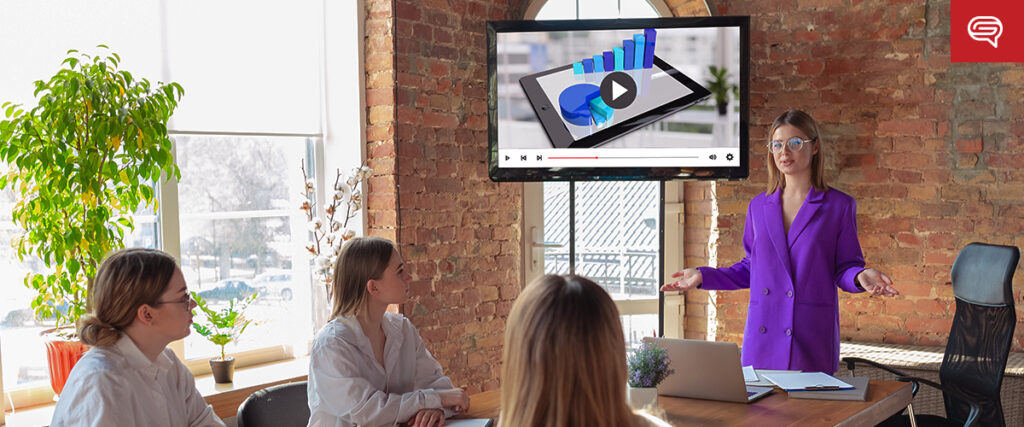 A woman in a purple blazer presents data on a wall-mounted screen to three people seated around a table in a modern office. The screen displays slides with graphs on a tablet. Sunlight streams in through large windows, casting a warm glow over the room. A potted plant is visible in the background.