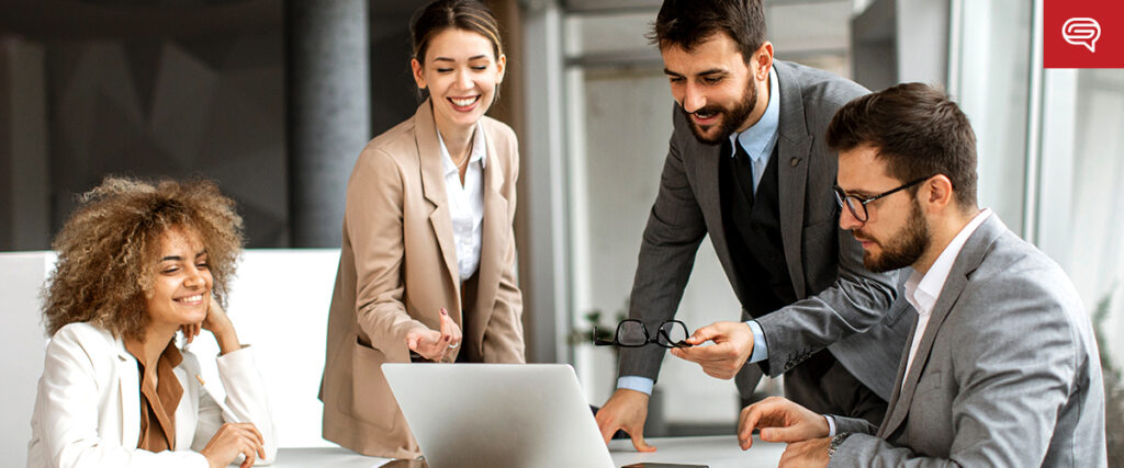 A diverse group of four business professionals are gathered around a laptop in a bright office. One person is sitting with a smile, while another, standing, is gesturing and looking at the screen. Two others are seated, attentively focusing on the pitch deck discussion.