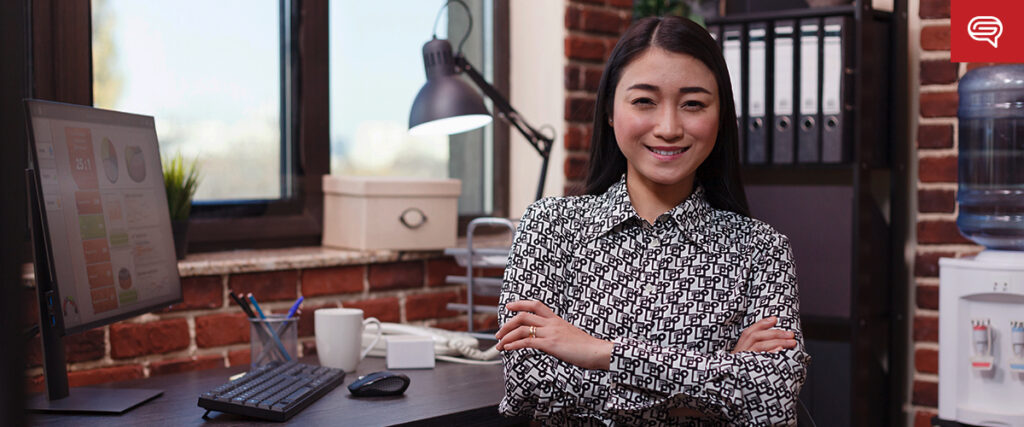 A woman with long dark hair stands confidently in an office, arms crossed and smiling. She wears a patterned blouse and stands next to a desk with a computer, lamp, and office supplies. The background features shelves with folders and a water cooler against a brick wall, possibly preparing her PowerPoint.