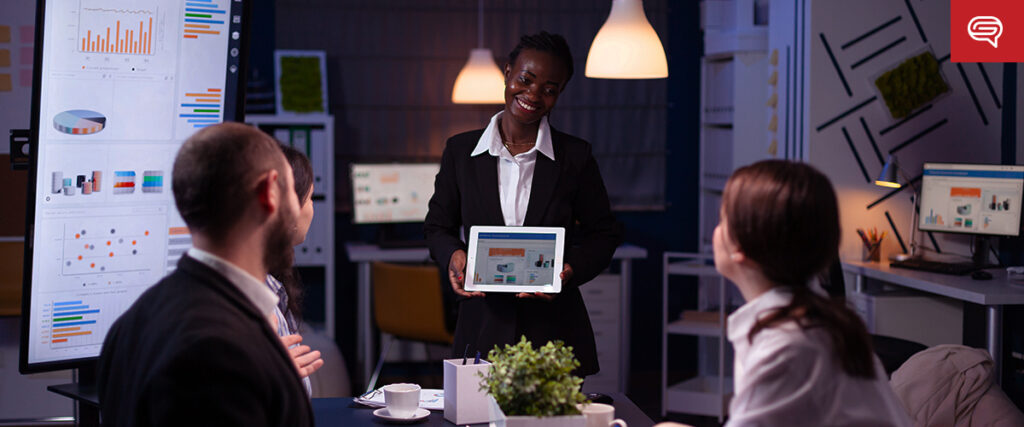 A person wearing a black blazer stands at the head of a conference table, holding a tablet displaying data charts. Three colleagues sit around the table, attentively listening to the presentation. Several monitors and charts with graphs are visible in the background.