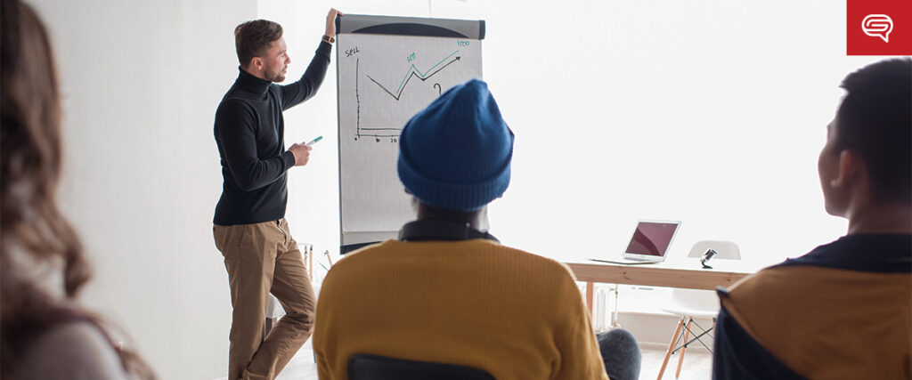 A man in a black turtleneck stands next to a flip chart with a slide, presenting to an audience. The audience, seated and casually dressed, focuses intently on the presentation. A laptop is placed on a table beside the man. The room is well-lit with natural light.