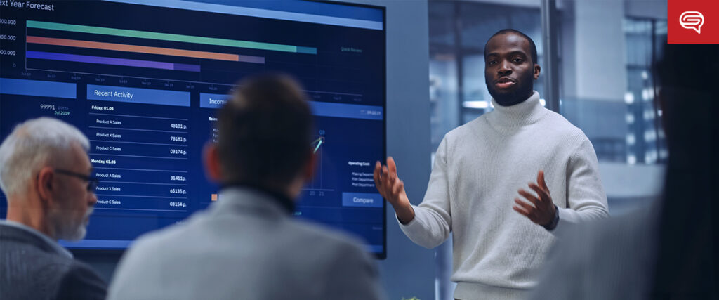 A man in a grey turtleneck shirt presents data on a large screen in a modern office setting. The screen displays various slides with graphs and charts. Three colleagues are seated facing him, attentively listening. The office has a professional and technology-driven atmosphere.