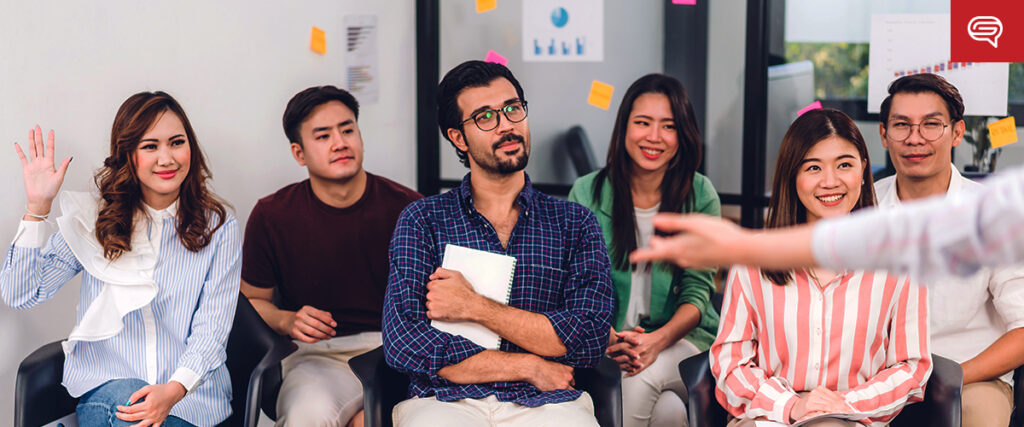 A diverse group of six people seated in a row, engaged in a discussion or meeting. One person is raising their hand, and another is holding a notebook. Colorful sticky notes are on the wall in the background. The focus is on a speaker presenting a pitch deck in the foreground.