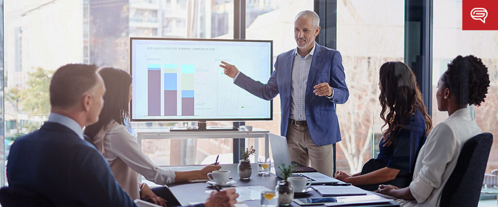 A man in a blue blazer presents a slide from his pitch deck displayed on a monitor to a group of five seated individuals in a modern conference room. The screen shows a bar graph and a line chart. Sunlight streams through large windows, providing a bright atmosphere.