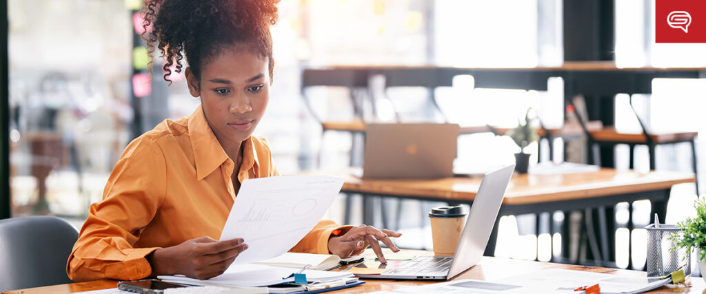 A woman with curly hair wearing an orange shirt is seated at a table, working with documents and a laptop. She is focused on a sheet of paper with graphs, possibly for her pitch deck. The table also has a coffee cup and various office supplies. The background shows a bright office space.