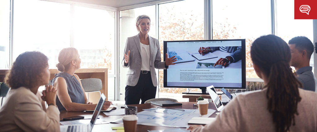 A group of people in a modern conference room attentively listens to a woman presenting. The presenter stands next to a large screen displaying a pitch deck. Laptops, notebooks, and coffee cups are on the table, indicating a professional business meeting.