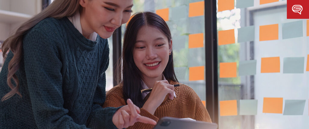 Two people collaborating on a project, one standing and pointing at a tablet held by the other who is sitting and smiling. The background shows a window covered with colorful sticky notes, possibly outlining their new PowerPoint slide template.