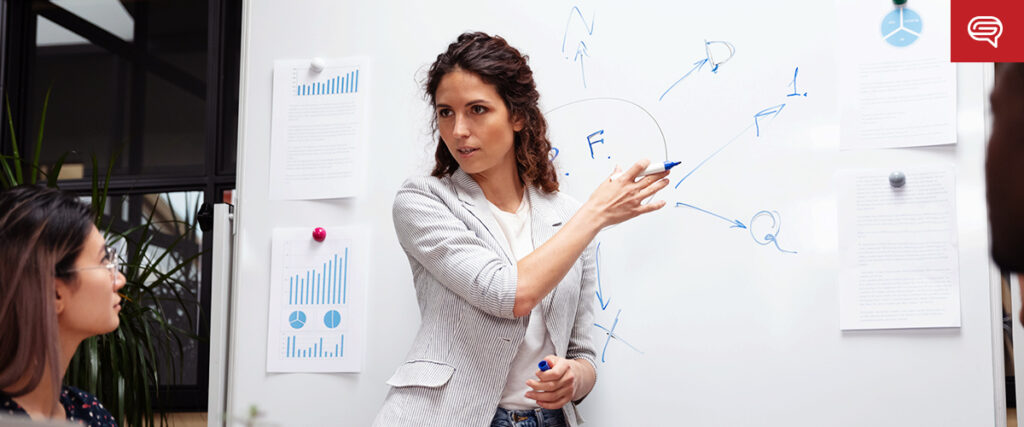 A woman stands at a whiteboard, explaining a concept with blue marker drawings to two colleagues. She is gesturing towards a diagram with various lines and letters, much like a slide in a PowerPoint presentation. Papers with charts and graphs are pinned to the whiteboard.