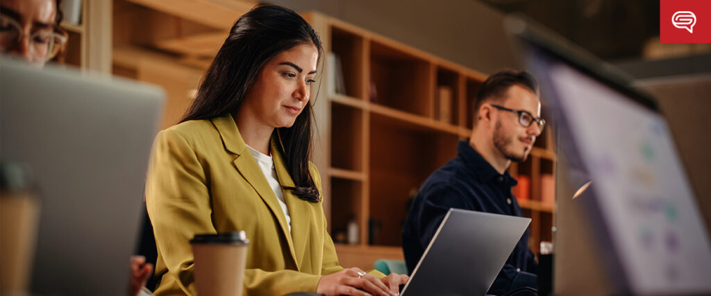 A woman in a yellow blazer and a man wearing glasses are working on laptops in an office. Coffee cups and shelves with books and other items are visible in the background, suggesting a focused and collaborative work environment as they prepare their PowerPoint presentation.